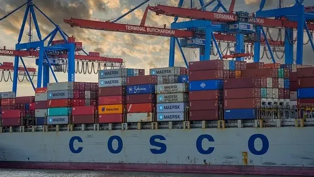 A large container ship is docked at a port, with cranes and cargo containers visible in the background.