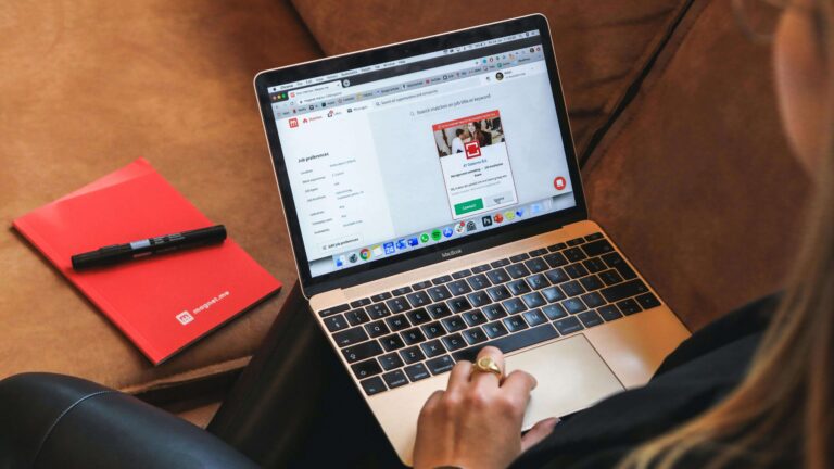 A woman sits on a couch, focused on her laptop, with a cozy living room setting in the background