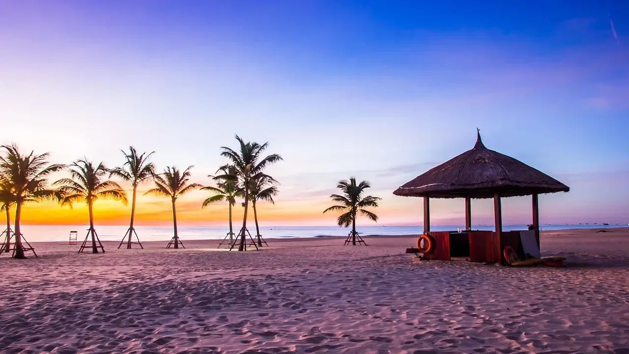 A serene beach at sunset featuring a hut and palm trees silhouetted against the colorful sky.