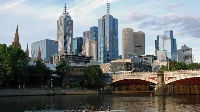 . A rowboat glides on the river, with tall buildings rising in the background against a clear sky