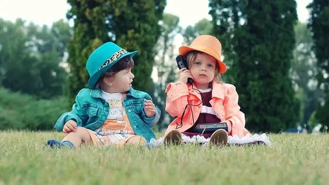 Two young children sitting on the grass, engaged in conversation while using a phone