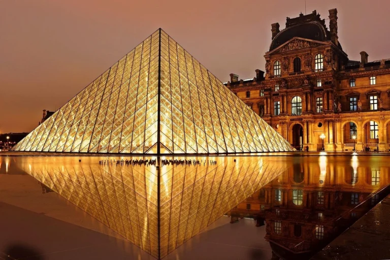 The Louvre illuminated at night, showcasing its iconic glass pyramid and historic architecture against a dark sky