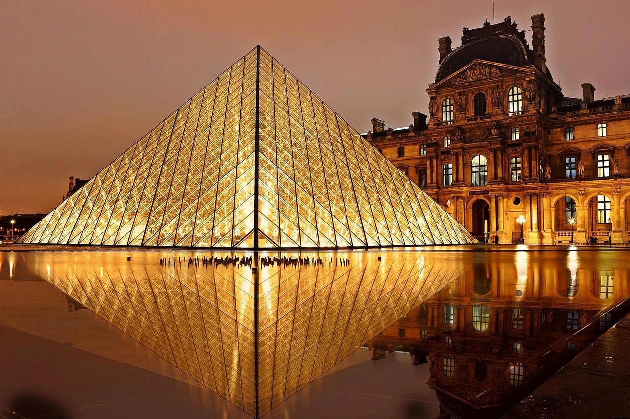The Louvre illuminated at night, showcasing its iconic glass pyramid and historic architecture against a dark sky