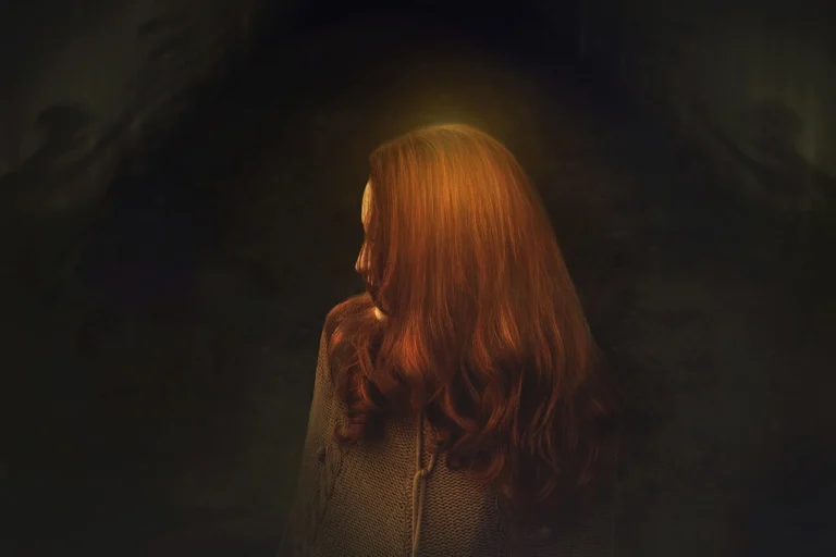 A woman with long red hair stands in a dark setting, her silhouette highlighted against the shadows around her